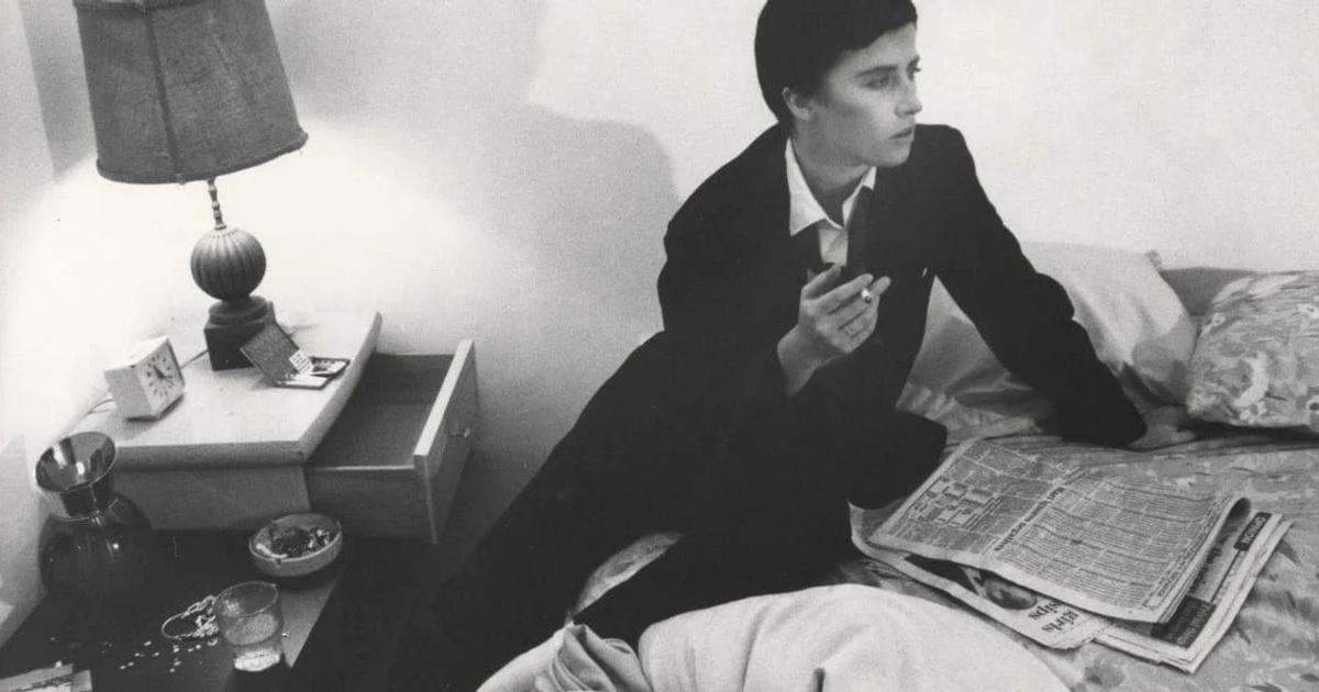 A black and white photograph of a young person in a dark coat sitting on a made bed with a newspaper spread open beside them, a lamp and a cluttered nightstand to one side.