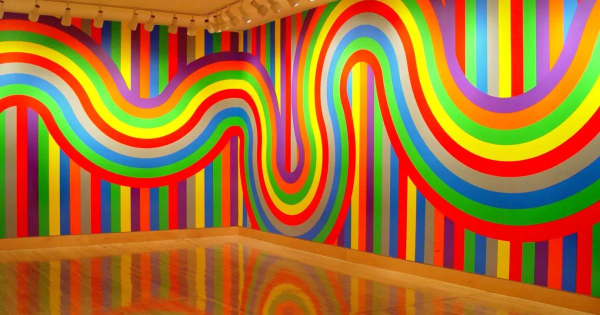 A gallery room painted with broad rainbow stripes that bend across the walls in a wave and continue over a reflective floor.