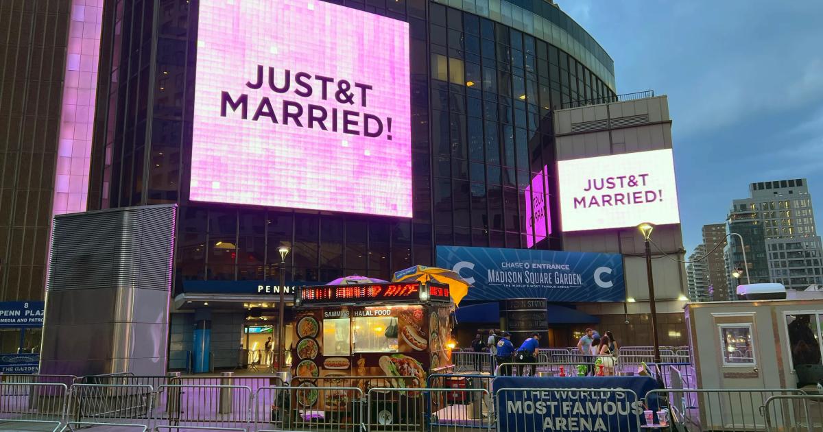 A city arena at dusk with two large illuminated screens reading Just Married, crowd barriers and passers by on the street in front.