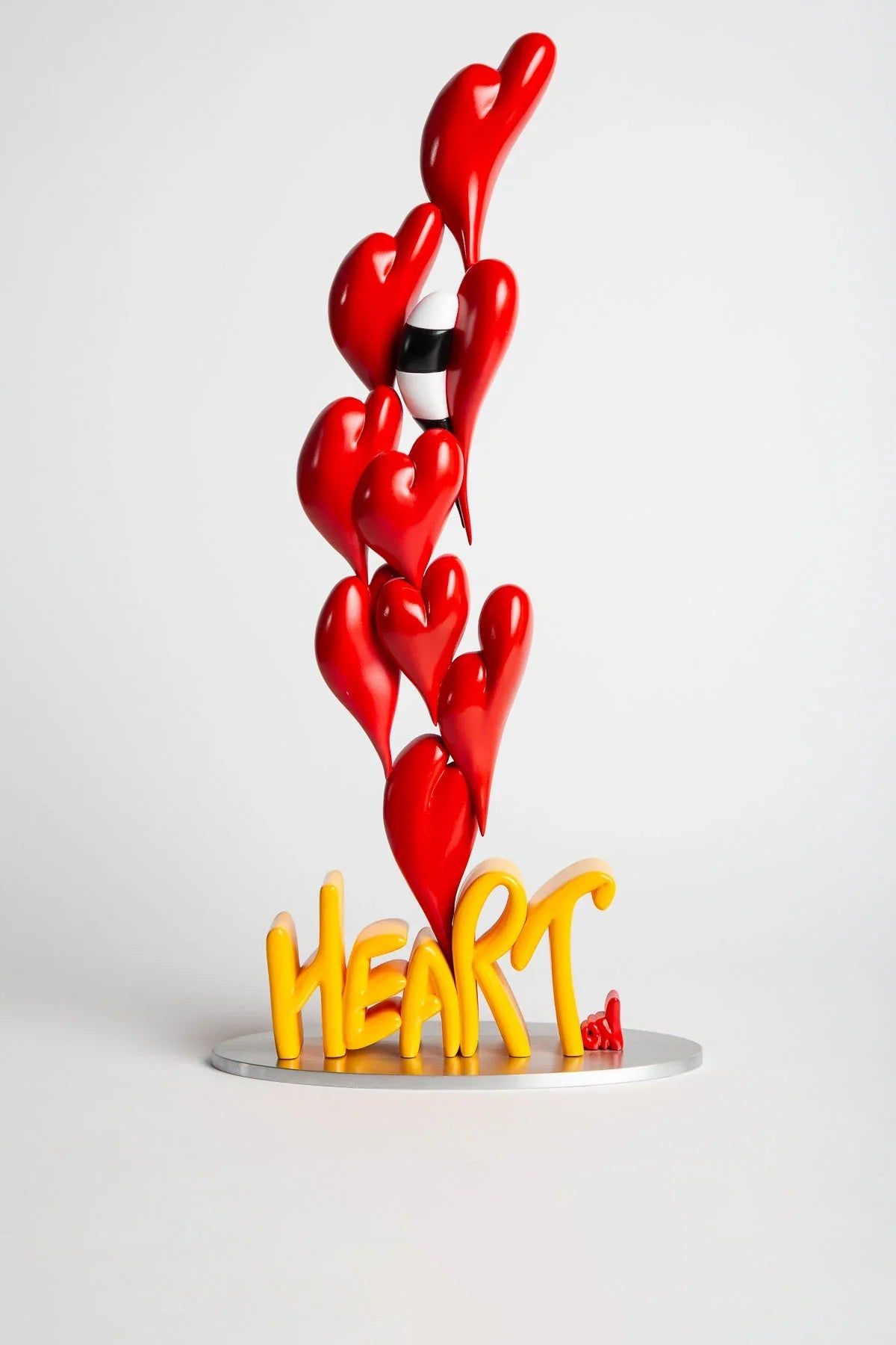 A resin sculpture of red hearts stacked into a column rising from the word HEART spelled in yellow on a circular metal base, photographed on white.
