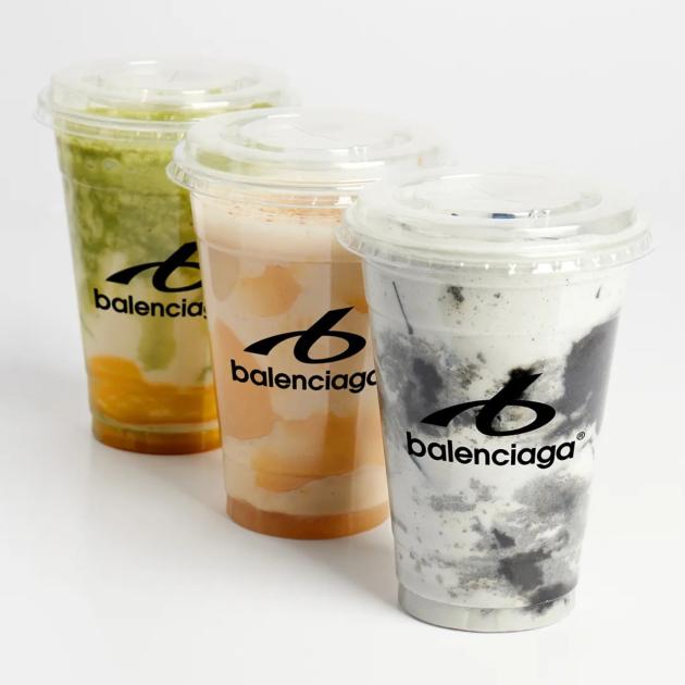 Three clear plastic cups of layered smoothie in green, orange and grey, each printed with a Balenciaga logo, on a white surface.