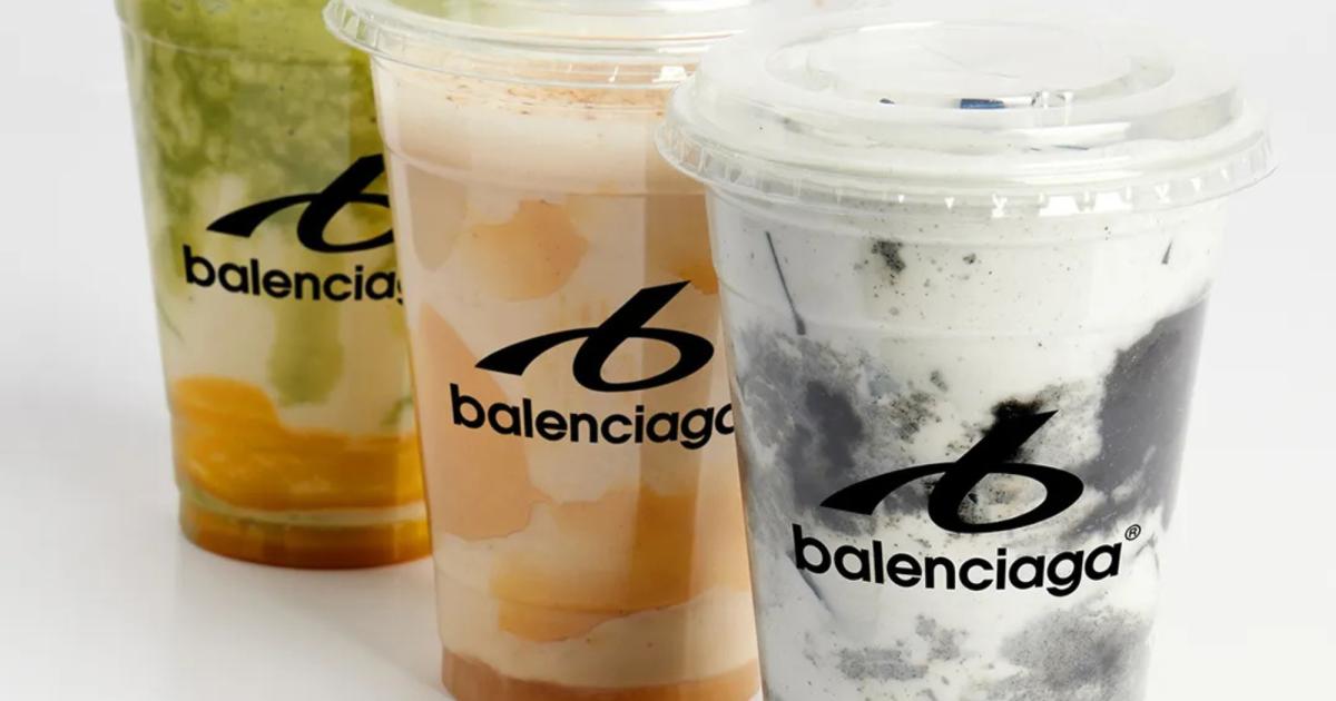 Three clear plastic cups of layered smoothie in green, orange and grey, each printed with a Balenciaga logo, on a white surface.