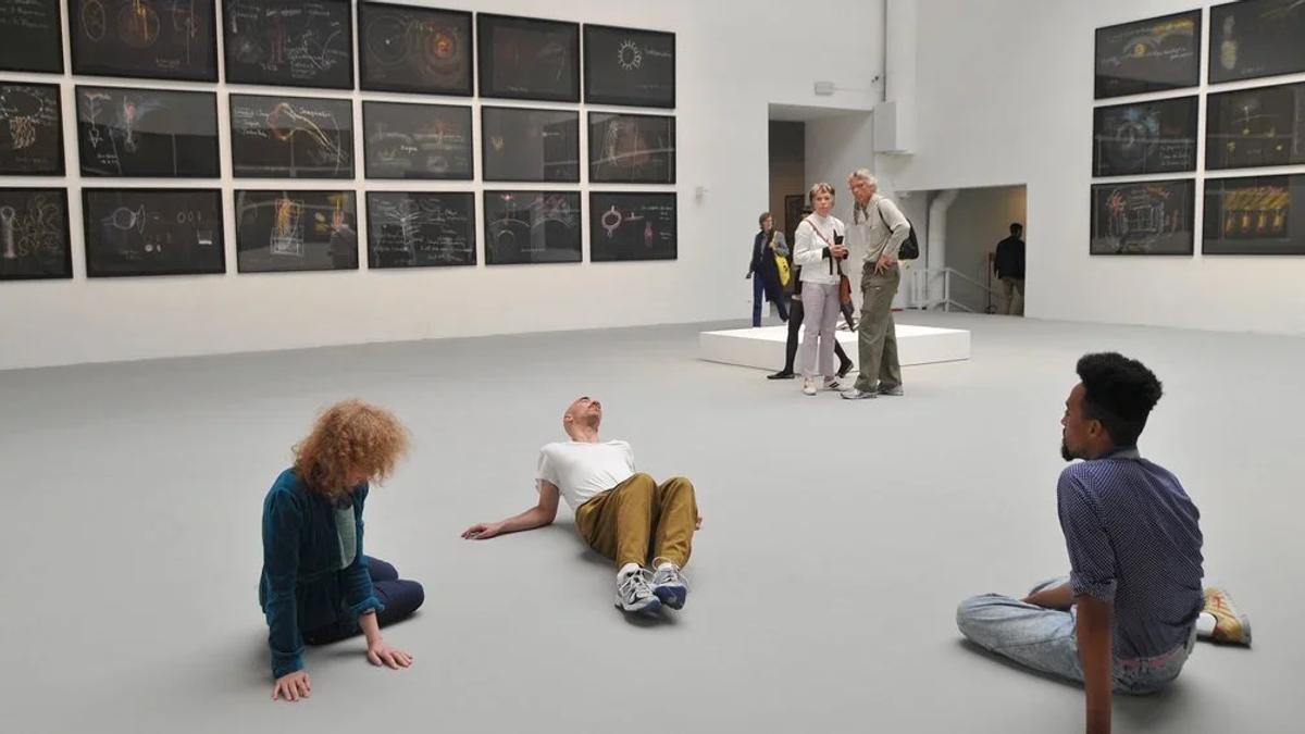 People sitting and lying on the floor of a bright gallery hung with rows of small framed works, a group of visitors standing at the far end.
