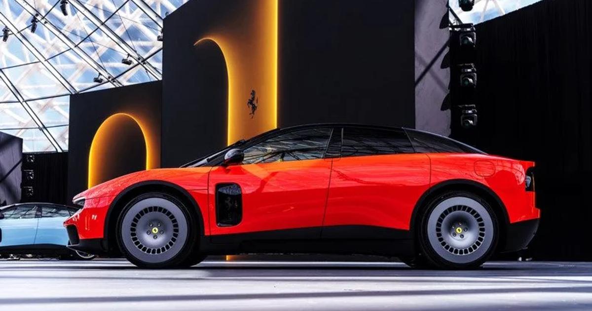 A red sports car photographed in profile on a show stand, a black backdrop behind it carrying a prancing horse emblem and glass roof structure above.