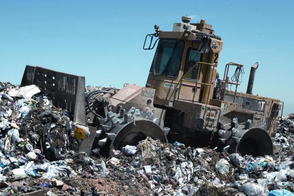 A heavy landfill compactor working across a spread of refuse at a waste site.