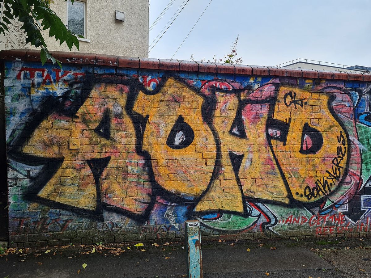 A brick wall in Bristol covered by layered spray-painted lettering, with traces of earlier red text still legible along the top and bottom edges. No people are in the frame.