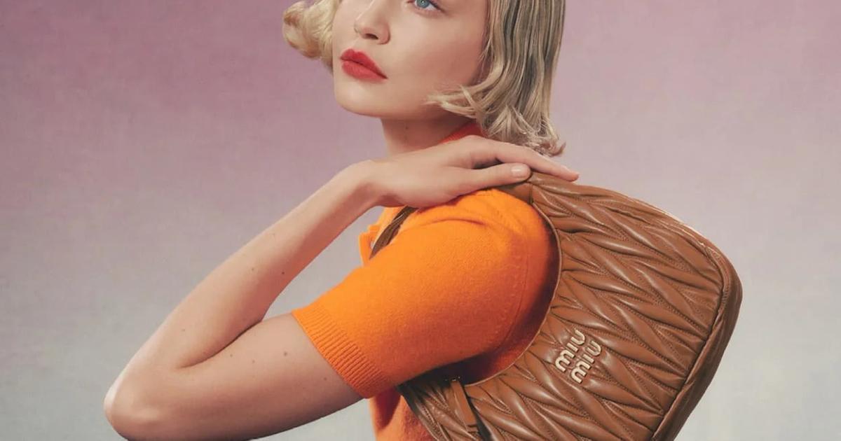 A blonde woman in a bright orange knit top holding a tan quilted Miu Miu shoulder bag, photographed against a soft pink backdrop.