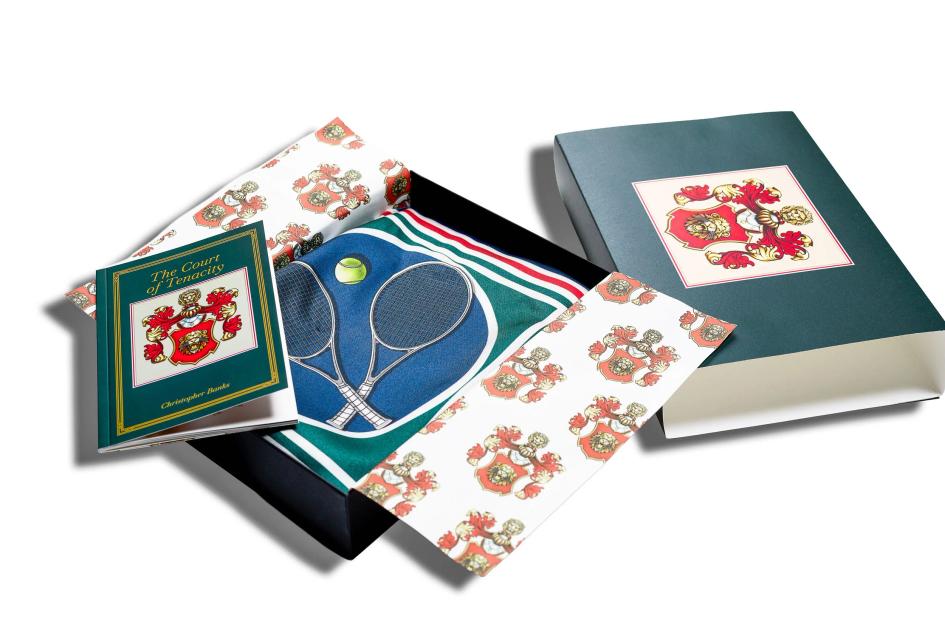 A boxed stationery set fanned out on a dark surface: cards and a cloth bound folder printed with a red and gold lion crest, and a green sheet showing crossed tennis rackets.