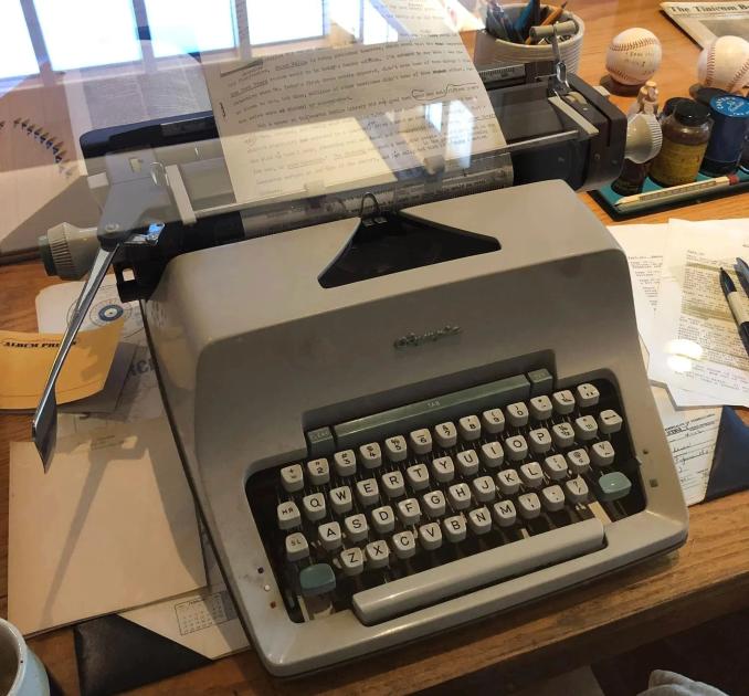 A manual typewriter on a wooden desk with a typed sheet in the platen, surrounded by loose handwritten pages, papers and baseballs on a shelf behind.