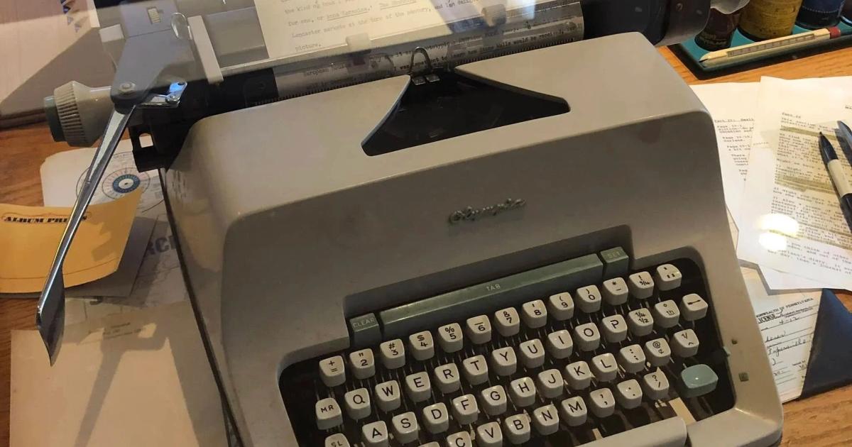 A manual typewriter on a wooden desk with a typed sheet in the platen, surrounded by loose handwritten pages, papers and baseballs on a shelf behind.