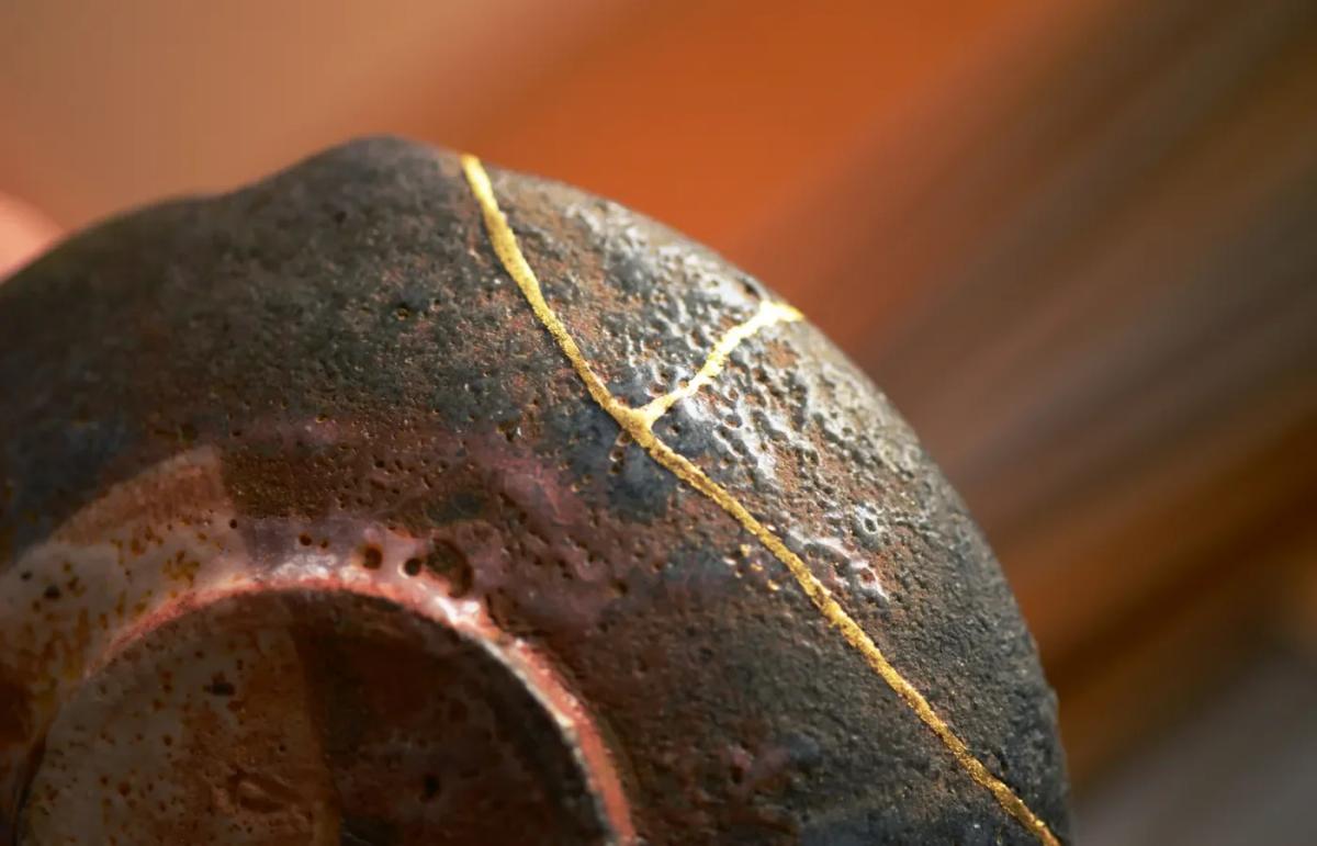 A close view of a rough dark ceramic bowl repaired with a fine gold seam running down its side and across the rim.