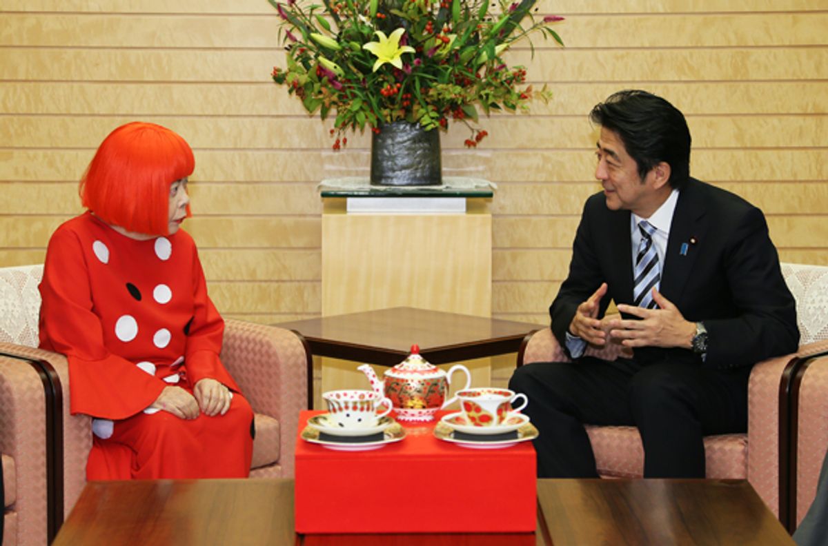 Yayoi Kusama, seated in a red wig and a red dress patterned with white dots, in conversation across a low red table.