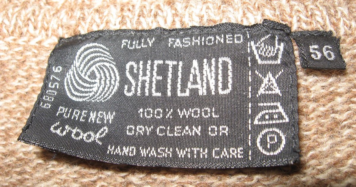 A sewn-in woven garment label reading 100 percent wool with care symbols, the object’s minimal testimony before the digital product passport.