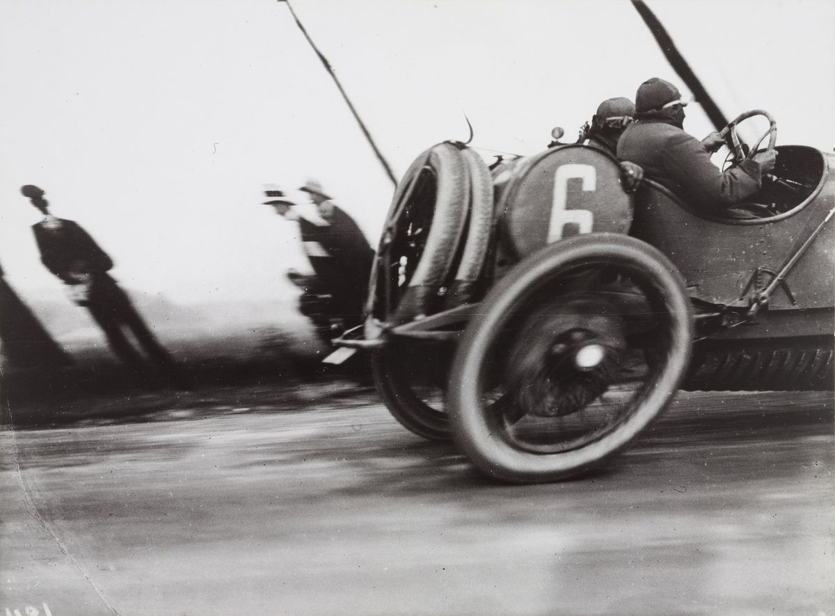 A black and white photograph of a racing car passing at speed, its wheels stretched into ovals by the camera's movement, with spectators leaning in a blur along the road.