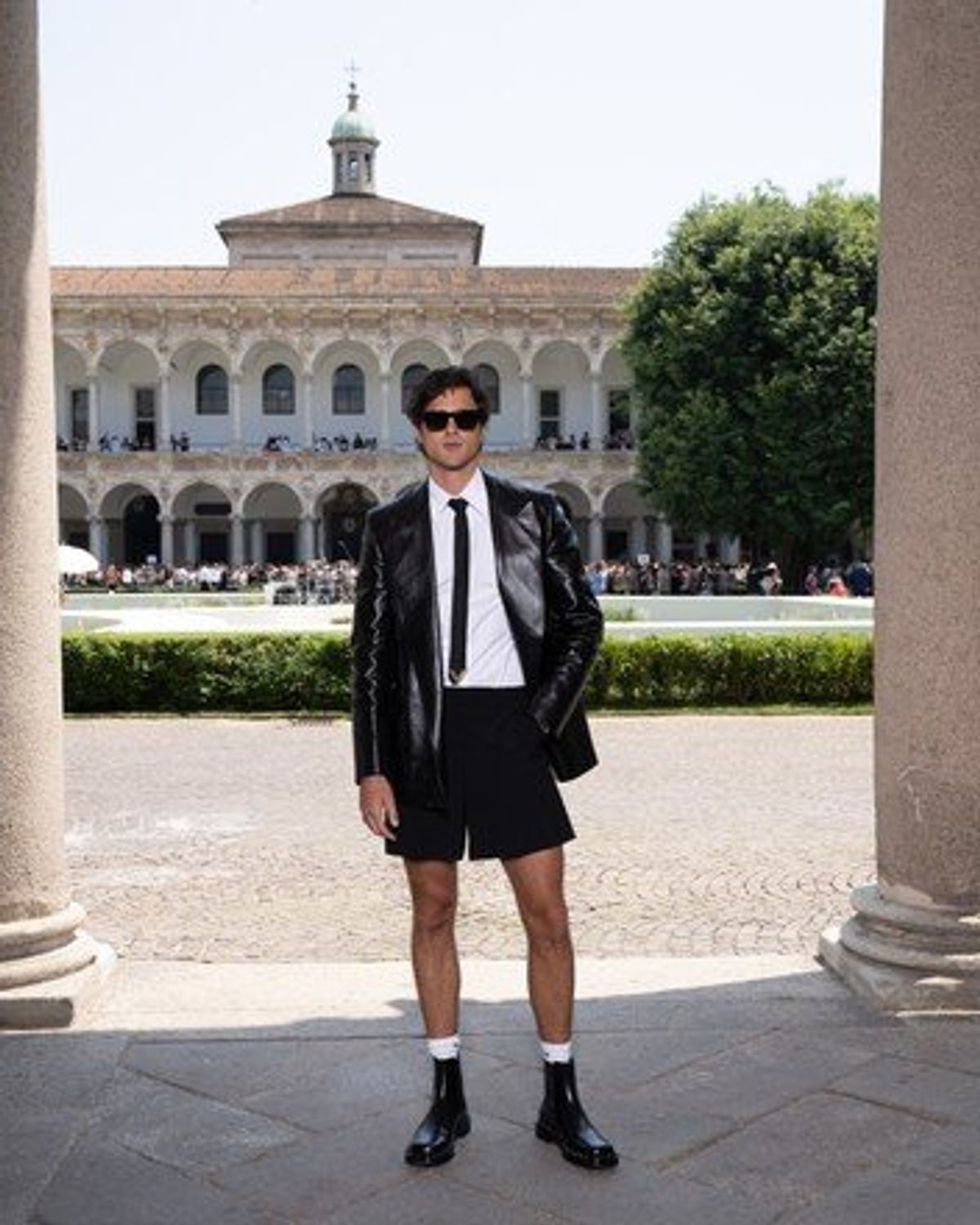 A man in a black leather jacket, white shirt, tie and shorts standing between stone columns in a sunlit university courtyard with arcaded buildings behind.
