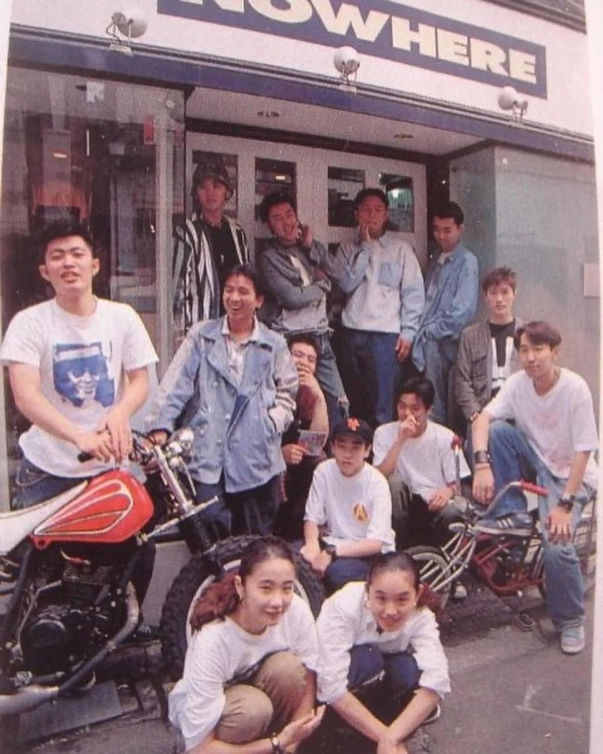 An archival photograph of a group of young people posed with motorbikes outside a shopfront, a sign reading Nowhere above the door.