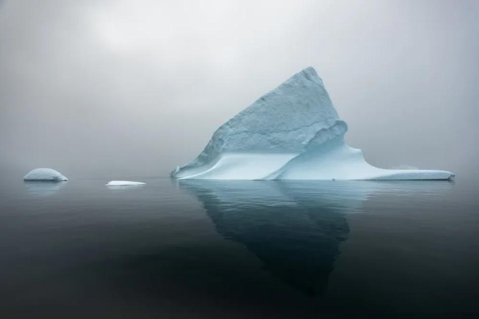 A single iceberg in flat grey water under heavy fog, its blue white peak reflected in the surface, with two small pieces of floating ice to the left.