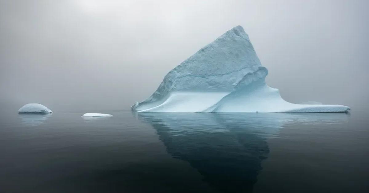 A single iceberg in flat grey water under heavy fog, its blue white peak reflected in the surface, with two small pieces of floating ice to the left.