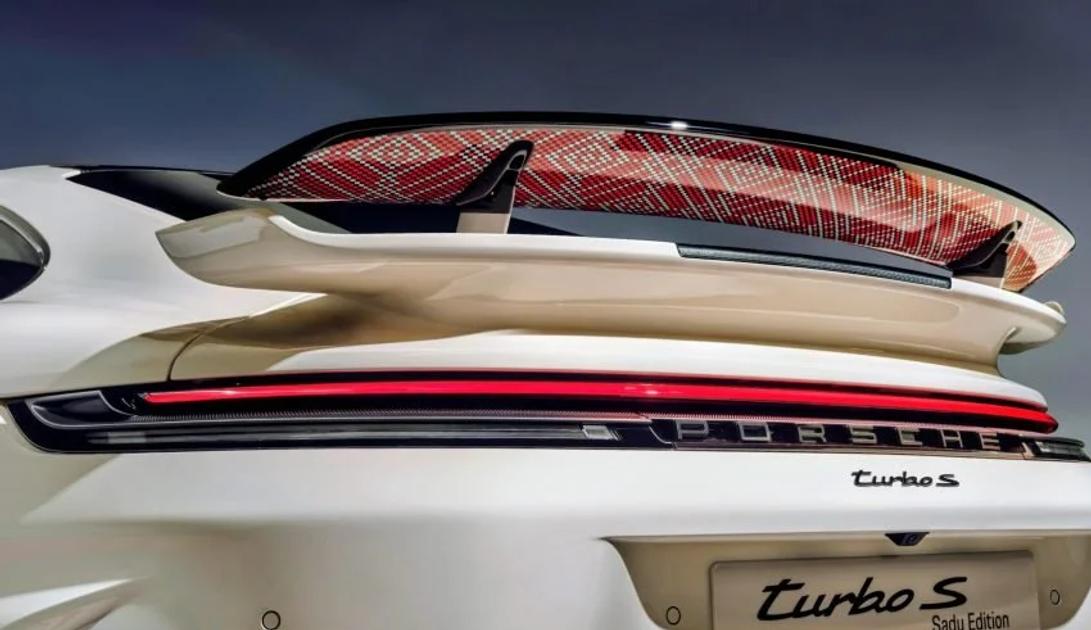 The rear of a white Porsche 911 Turbo S, its raised wing carrying an intricate red and gold woven pattern on the underside, with Turbo S and Sadu Edition lettering below.