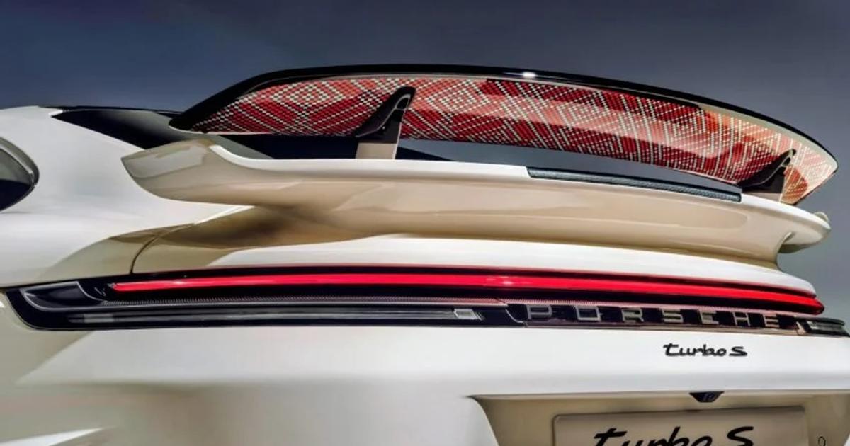 The rear of a white Porsche 911 Turbo S, its raised wing carrying an intricate red and gold woven pattern on the underside, with Turbo S and Sadu Edition lettering below.