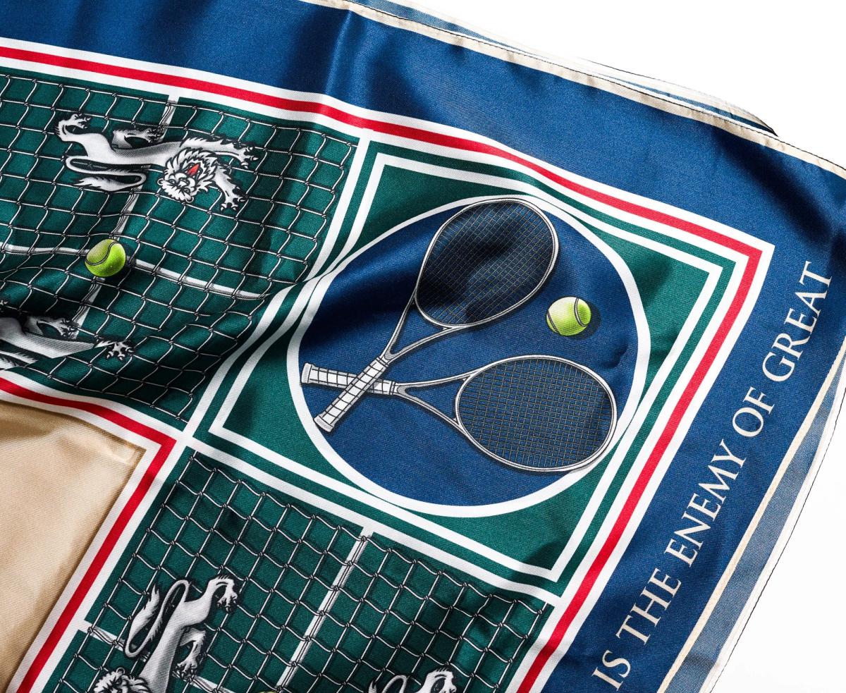 A detail of the scarf, crossed tennis rackets and a ball inside a navy roundel on a green court ground, with the words is the enemy of great along the border.