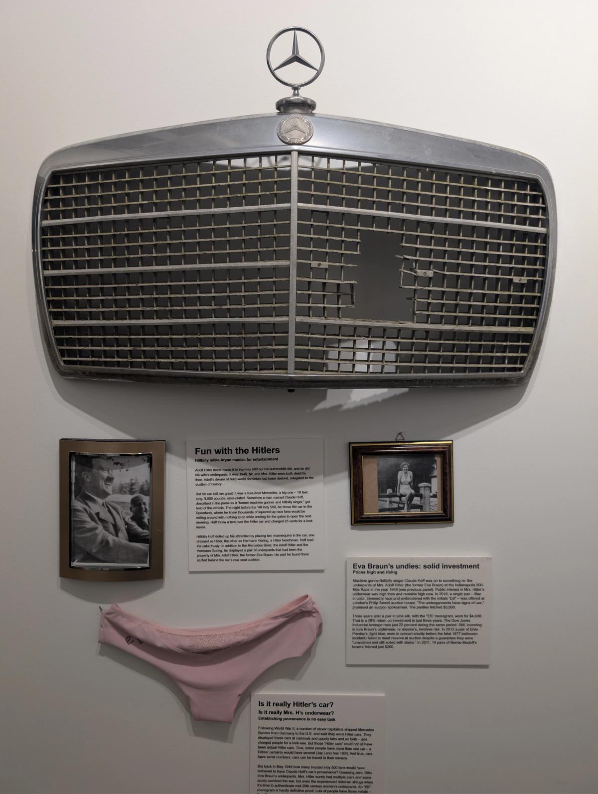 A gallery wall carrying a large Mercedes radiator grille with the three-pointed star mounted above framed photographs, printed wall texts, and a pair of pink underpants displayed flat.