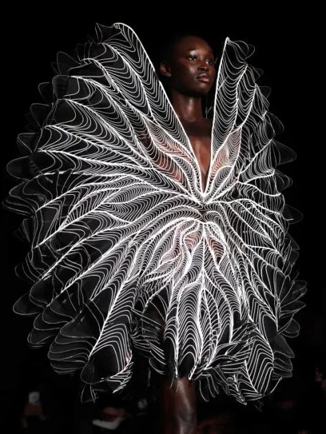 A model on a darkened runway wearing a large circular dress built from fine white filaments that radiate from the centre of the body in a symmetrical wing like pattern.