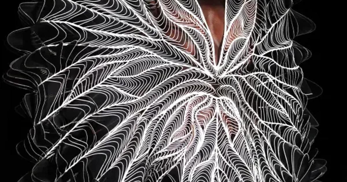 A model on a darkened runway wearing a large circular dress built from fine white filaments that radiate from the centre of the body in a symmetrical wing like pattern.