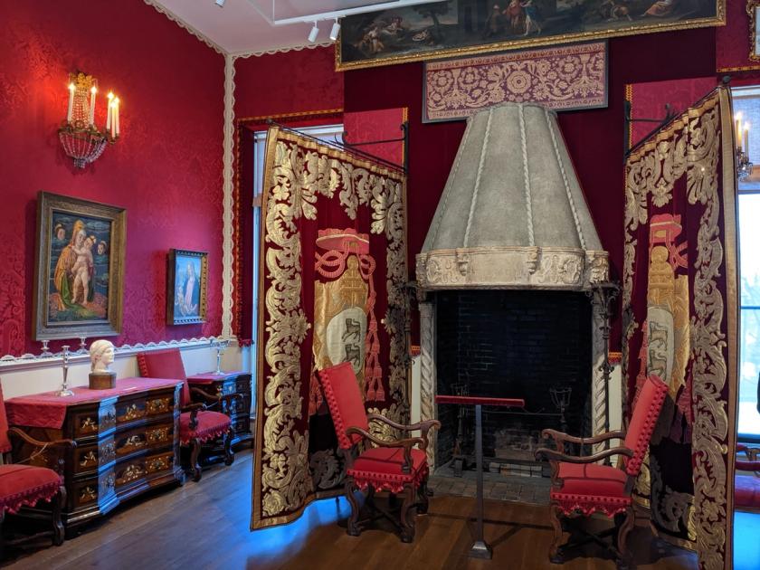 An interior gallery at the Isabella Stewart Gardner Museum, hung densely with paintings and tapestries above patterned walls and antique furniture, arranged as a domestic room rather than a modern gallery.