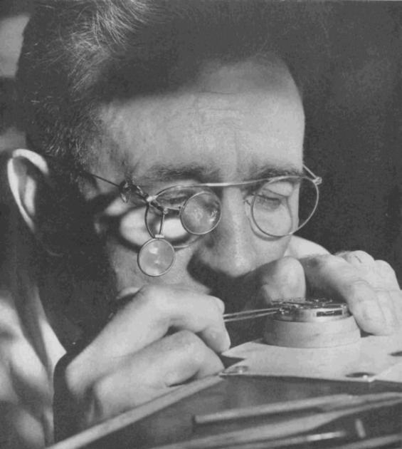 A watchmaker seated at a bench, loupe to one eye, working on the movement of a watch held in one hand, surrounded by tools and small parts.