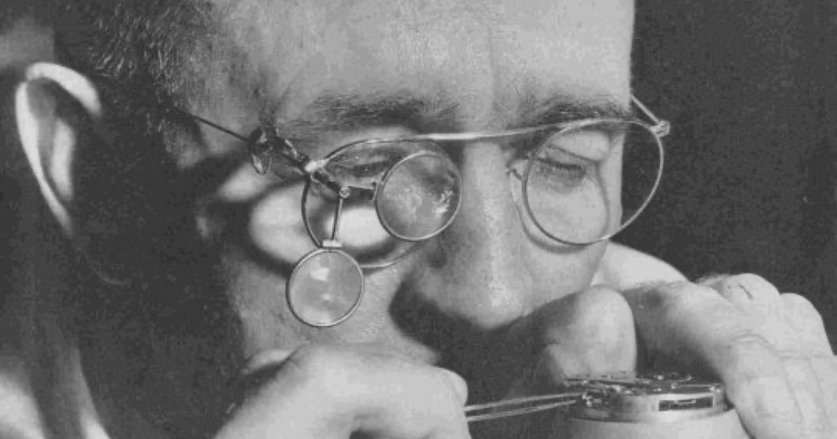 A watchmaker seated at a bench, loupe to one eye, working on the movement of a watch held in one hand, surrounded by tools and small parts.