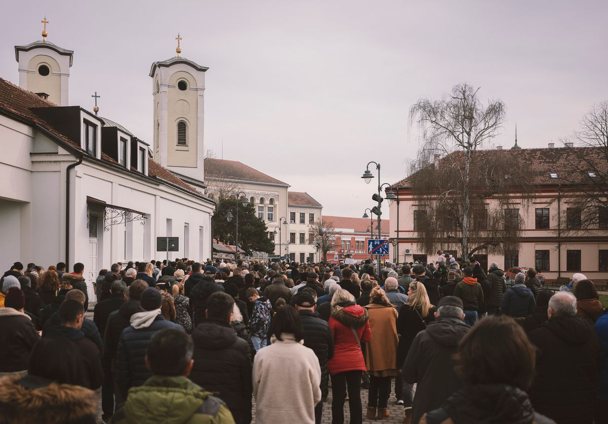 A dense winter crowd filling a Serbian town square, seen from behind, with the twin towers of an orthodox church rising above the rooftops under a grey sky.