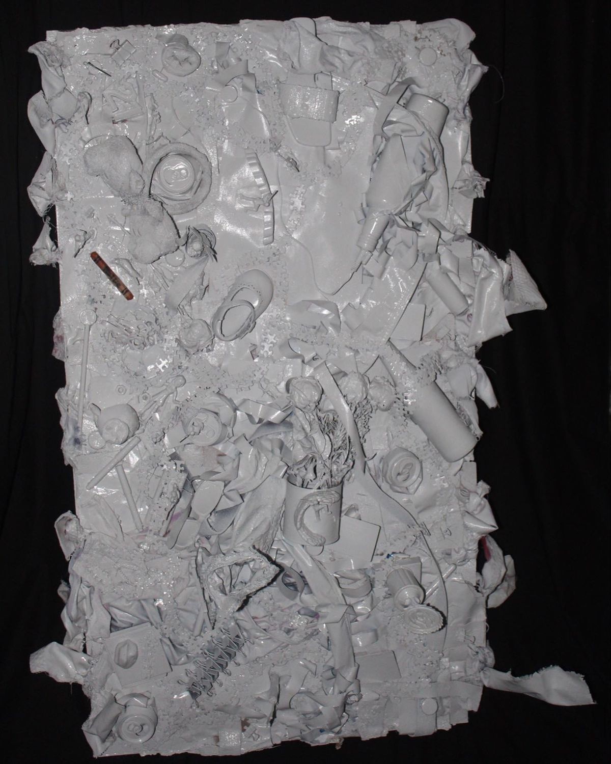A dense white relief panel. Packaging, caps, lids, doll clothes, figurines, puzzle pieces, a flag and desk supplies are massed together and painted a single uniform color. Near the upper left one crayon remains unpainted, faded orange, with handwriting on its wrapper.