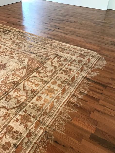 A Persian carpet pattern carved into a red oak floor, seen at an angle. The field, borders and floral medallions are cut in relief, the fringe is rendered as individual strands lying in different directions, and the surrounding gallery floor continues as ordinary polished oak.
