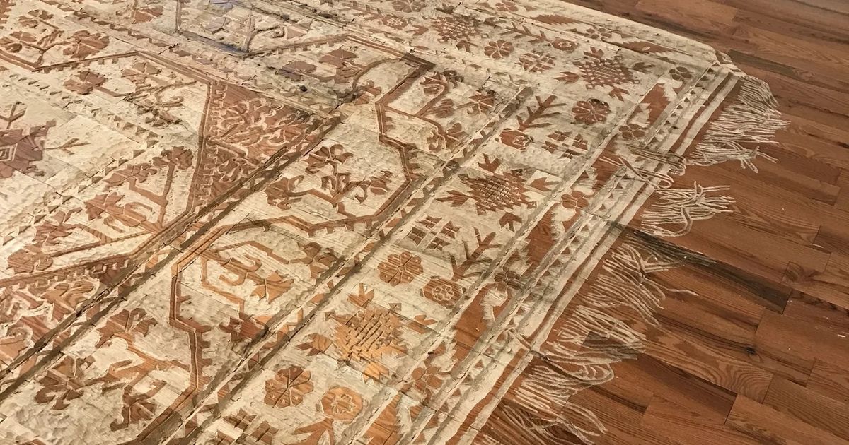 A Persian carpet pattern carved into a red oak floor, seen at an angle. The field, borders and floral medallions are cut in relief, the fringe is rendered as individual strands lying in different directions, and the surrounding gallery floor continues as ordinary polished oak.