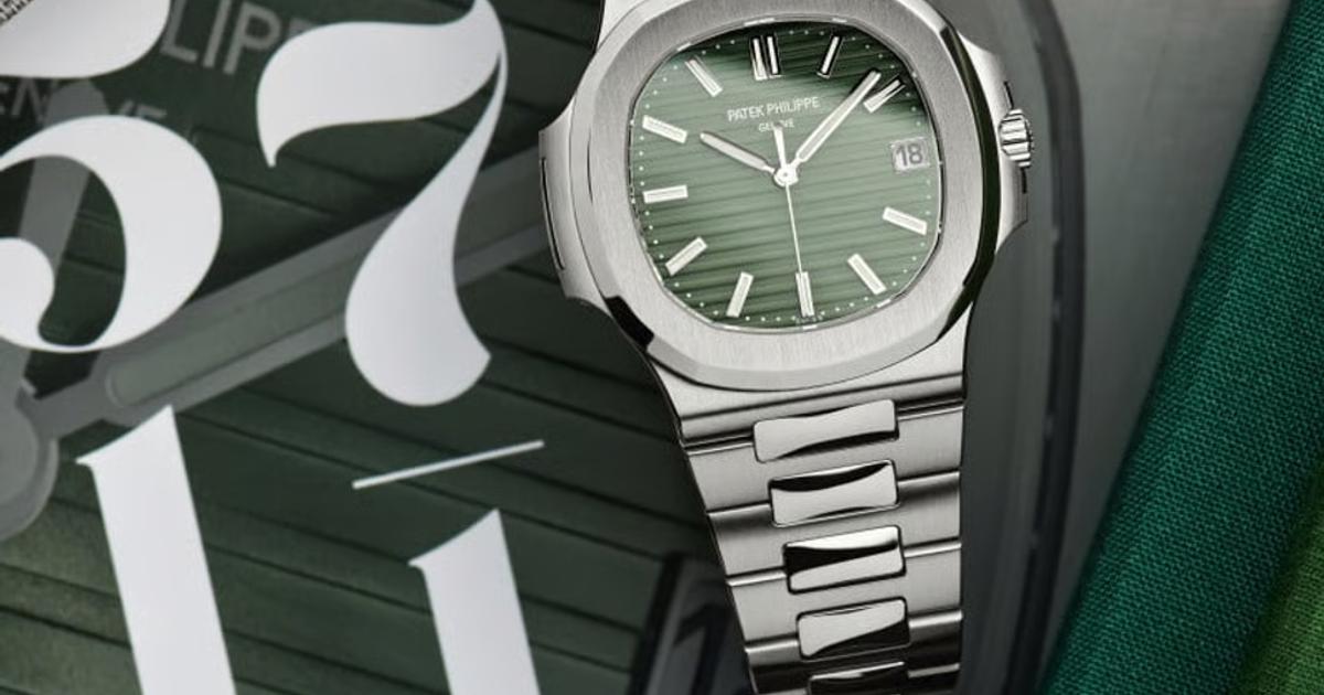 A steel sports watch with a green textured dial and integrated bracelet, resting on fabric in front of a large printed number.