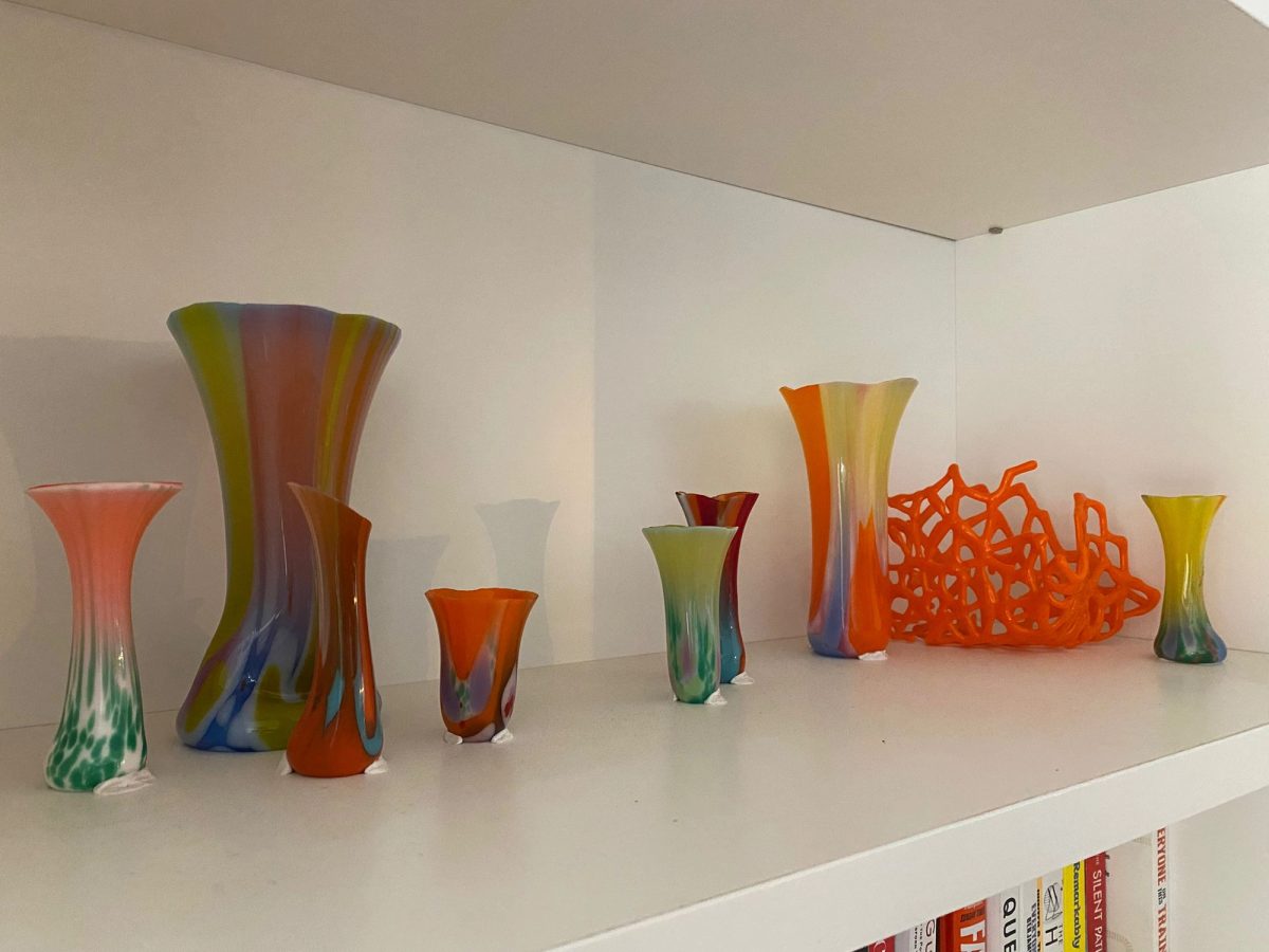 A wide shelf display of multiple colorful glass vessels and an orange lattice form arranged across a white built-in shelf.
