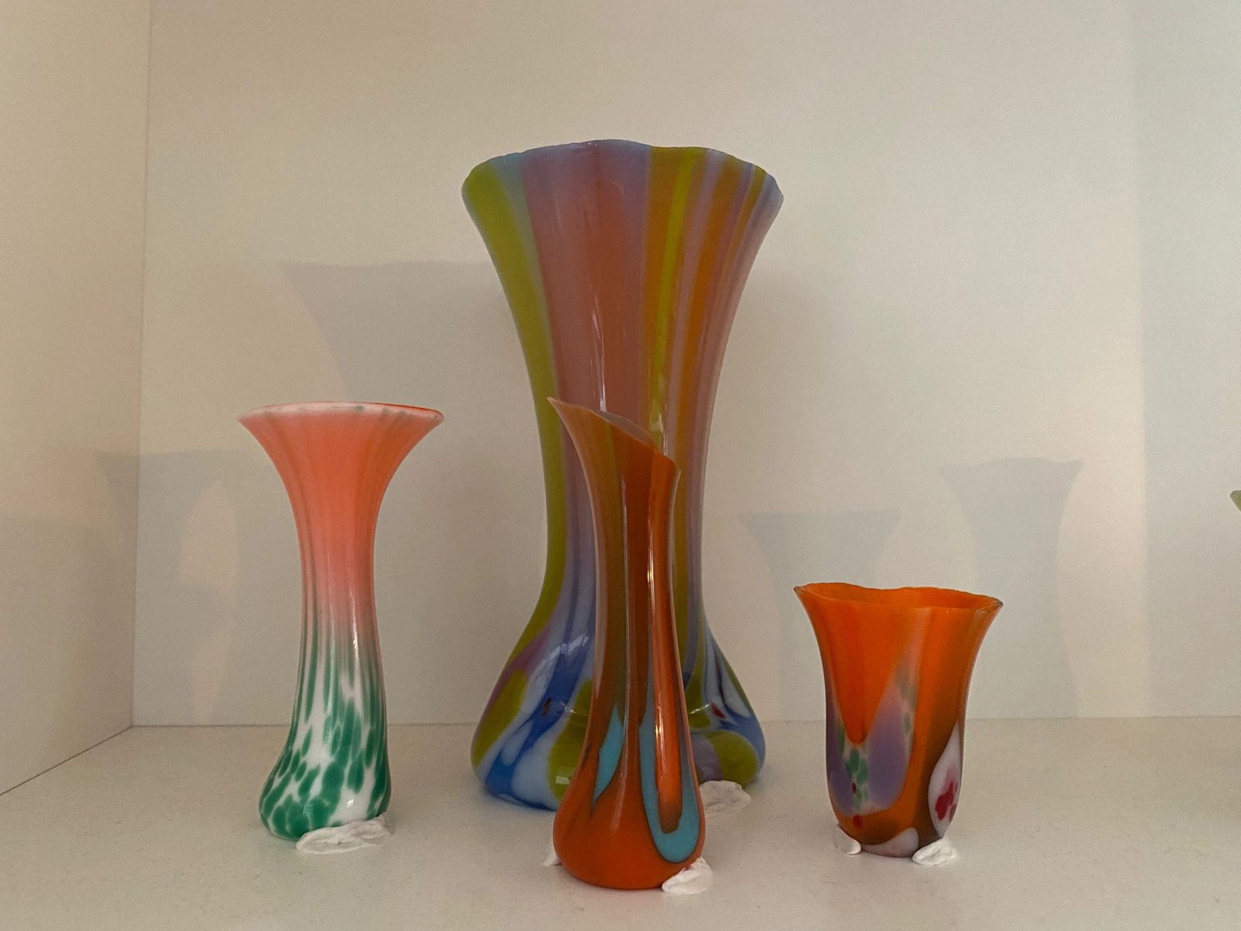 Four colorful hand-formed glass vessels displayed on a white shelf, including pink, orange, blue, and green marbled forms in different heights.