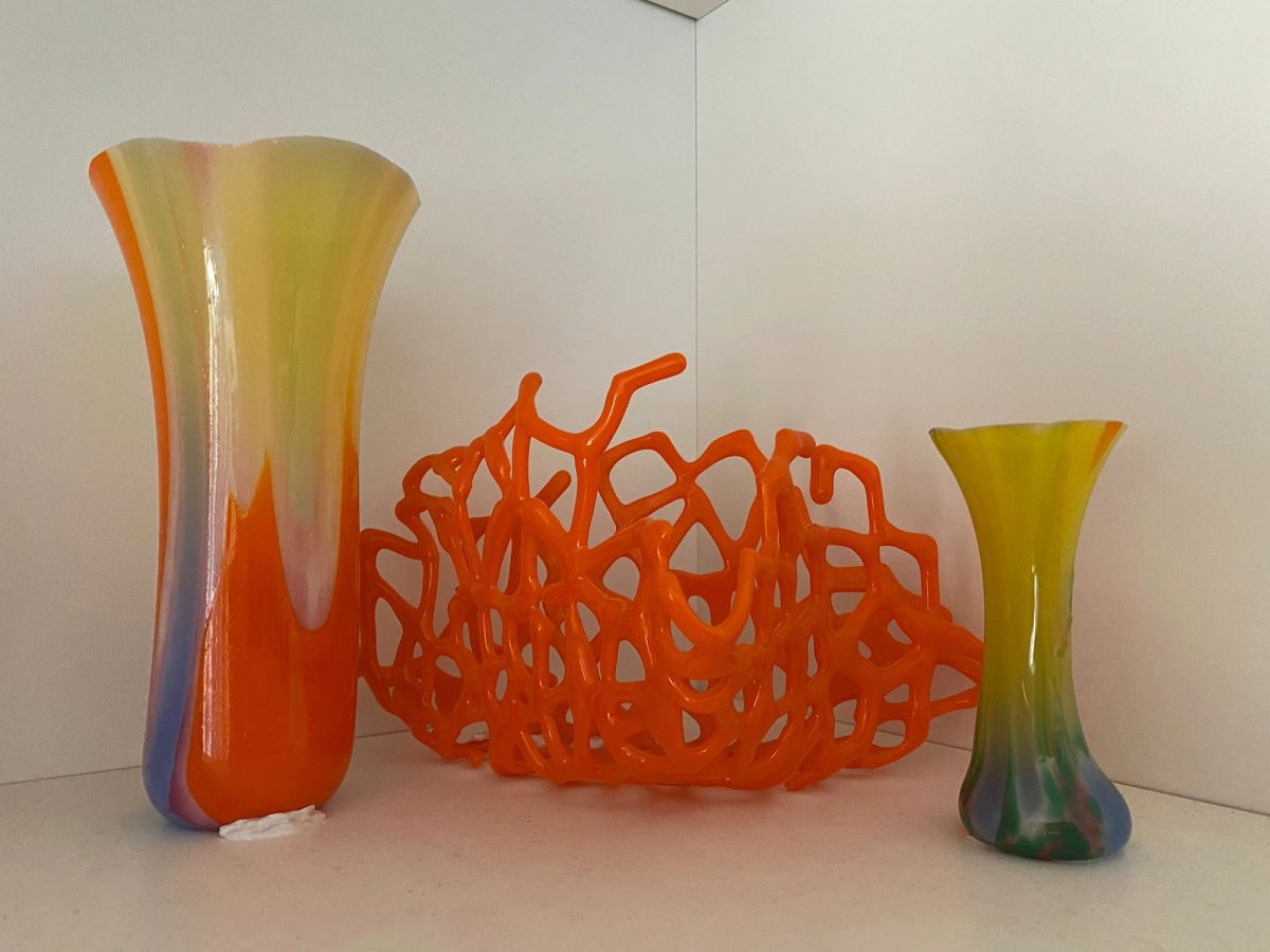 An orange web-like glass form displayed between two tall translucent vessels on a white shelf.