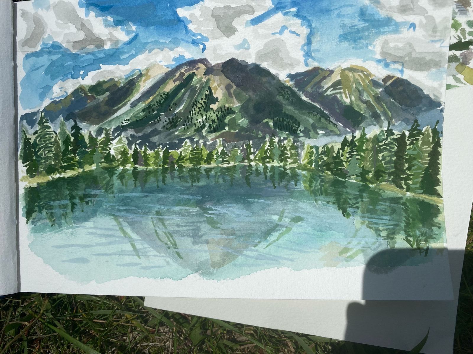 A watercolor landscape showing mountains, a lake, pine trees, and blue-grey clouds painted in loose layered washes.