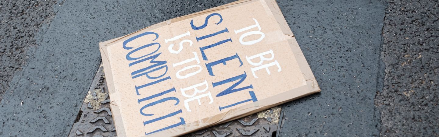 a placard lies on the street with the words 'to be silent is to be complicit' | near and far enemies of fierce compassion | tempo therapy and consulting