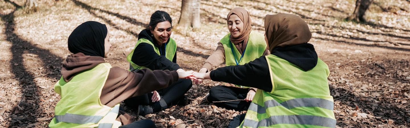 four women wearing head scarves and high visability jackets sit in a circle, each reaching a hand into the centre  | collective care | Tempo Therapy and Consulting