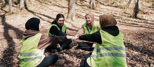four women wearing head scarves and high visability jackets sit in a circle, each reaching a hand into the centre  | collective care | Tempo Therapy and Consulting