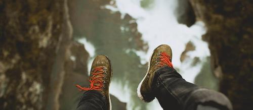 closeup of legs dangling over a precipice with jeans and hiking boots in the foreground and a long drop to rocks below