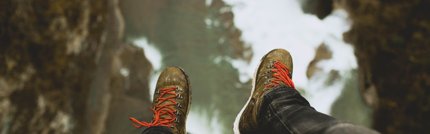 closeup of legs dangling over a precipice with jeans and hiking boots in the foreground and a long drop to rocks below