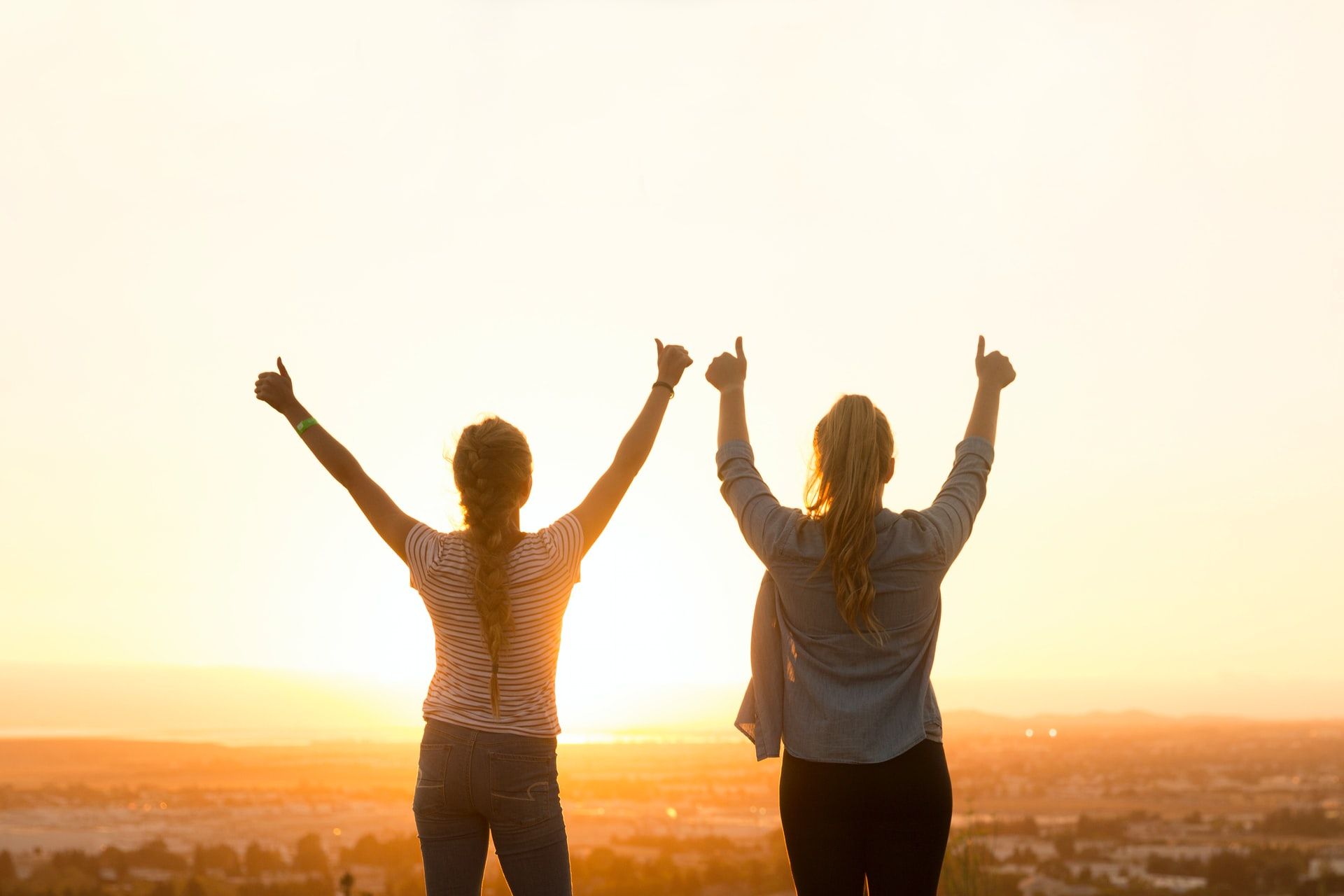 two women stand on a hill with their arms in the air looking out across a sunrise