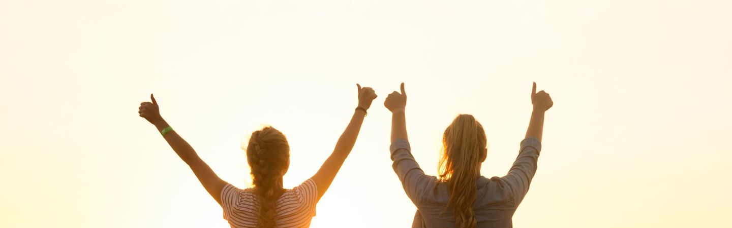 two women stand on a hill with their arms in the air looking out across a sunrise