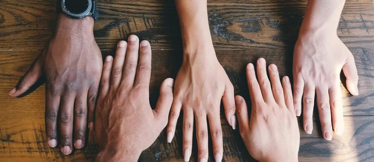 5 hands with varying shades of brown and white skin lay flat on a wooden desk | group supervision | Tempo Therapy and Consulting