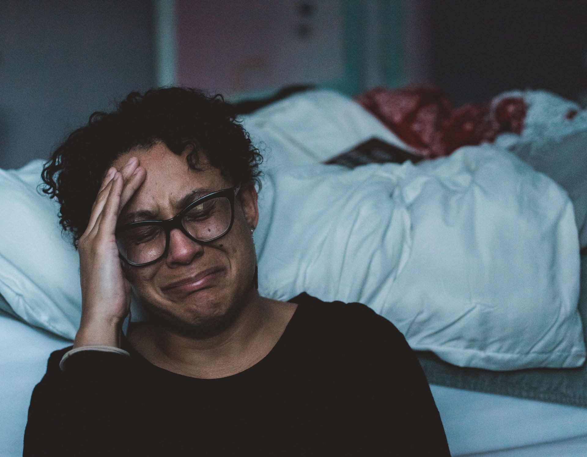 A woman with  glasses, brown skin and short black curly hair sits in front of a white unmade bed crying | burnout symptoms | Tempo Therapy and Consulting