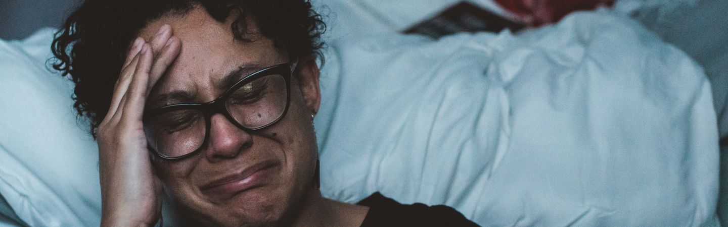 A woman with  glasses, brown skin and short black curly hair sits in front of a white unmade bed crying | burnout symptoms | Tempo Therapy and Consulting